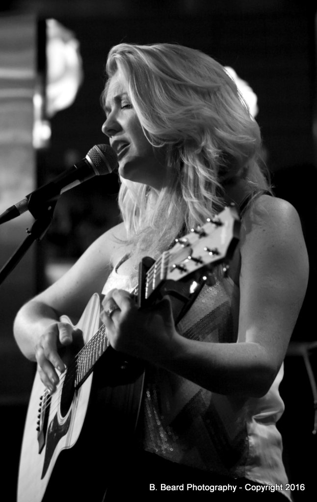 Blonde woman holding a guitar, on a stage.